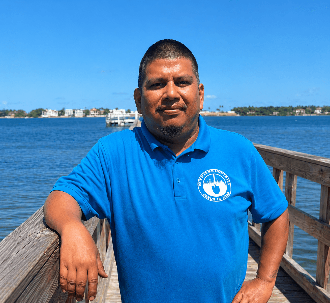 Man in a blue polo shirt leaning on a wooden pier railing by the water.