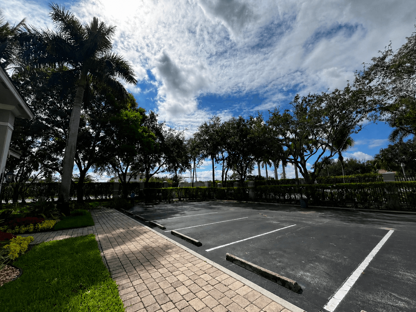 Empty asphalt parking lot with brick walkway, surrounded by lush trees under a cloudy sky.