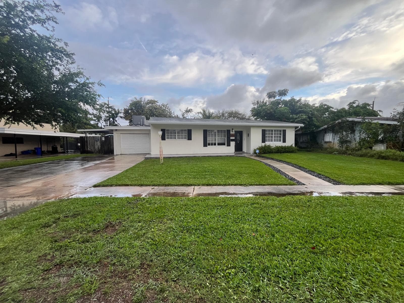 White single-story house with black shutters, a green lawn, and a wet concrete driveway.
