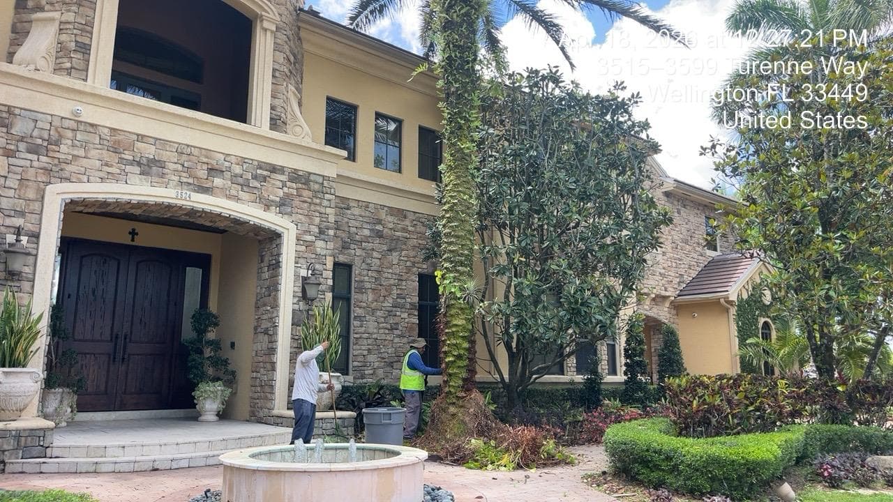 Two workers maintain the landscaping of a large stone house with a front fountain.