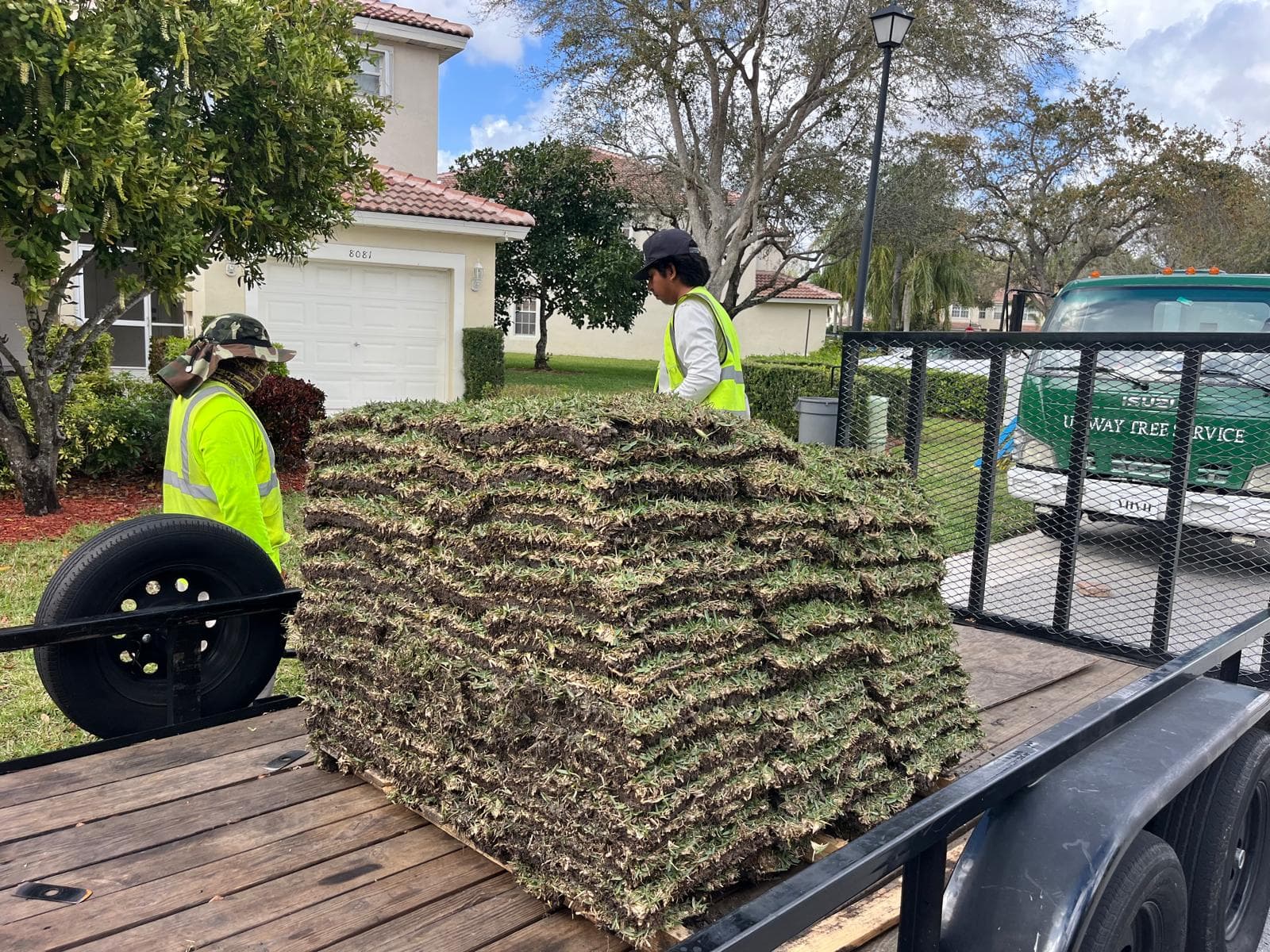 Two workers in safety vests stand by a large stack of sod on a trailer.