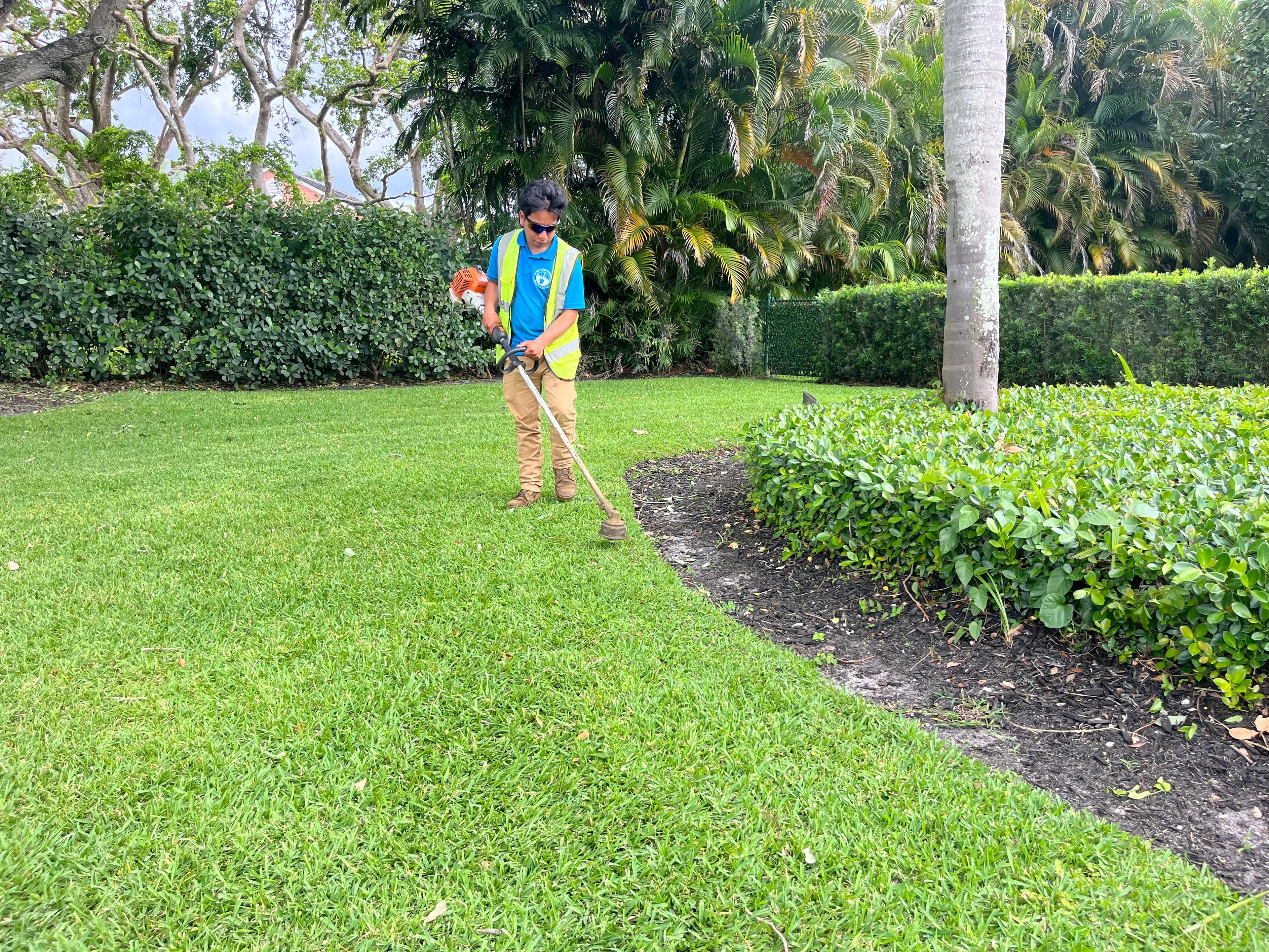 Landscaper in a safety vest uses a string trimmer to edge a lush green lawn.