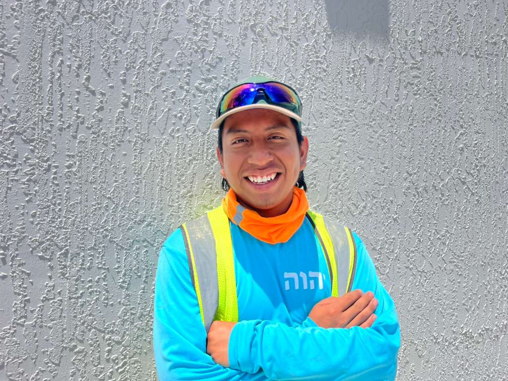 Smiling man in a blue shirt and safety vest against a textured gray wall.